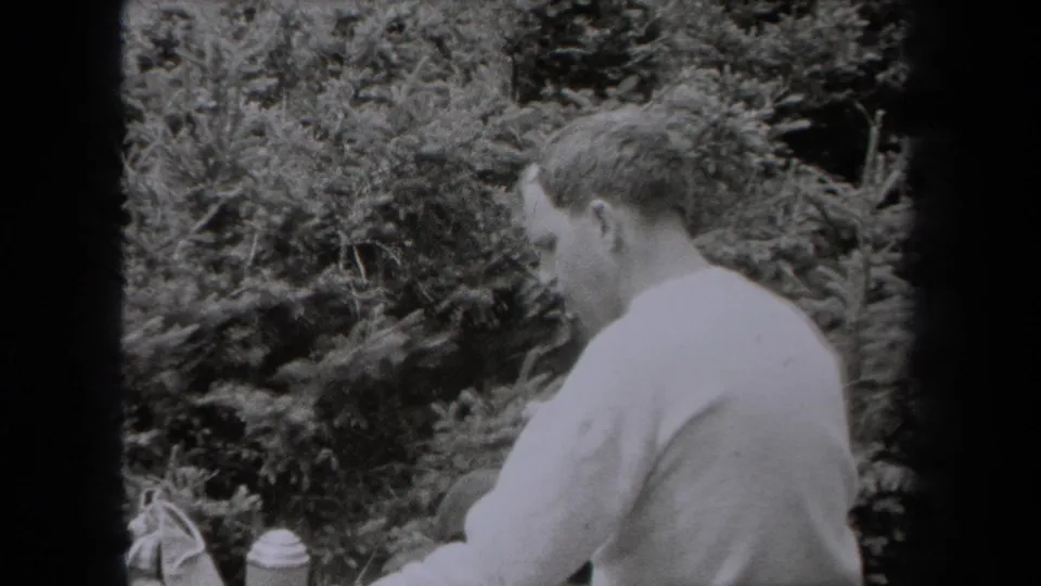 In this 1938 New York 8mm frame, a man in a light shirt sits outdoors among evergreen trees with a small container nearby.