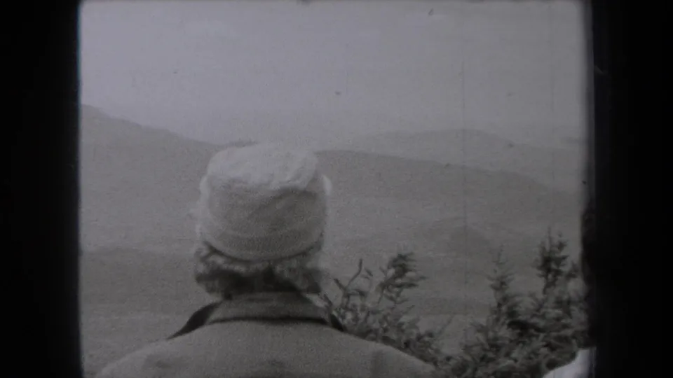 Adult wearing a soft cap and jacket, seen from behind in 1938 New York footage, facing a hazy hillside view.