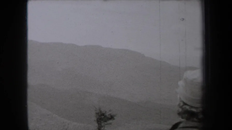 1938 New York 8mm frame with a hat-wearing person at the right edge looking toward pale, wind-hazed dunes.