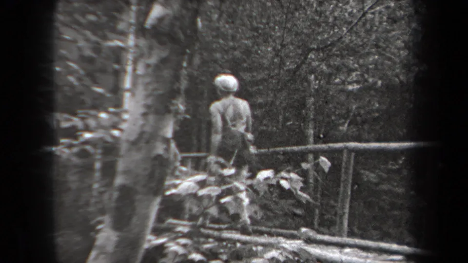 1938 New York 8mm frame showing a woman from behind on a rustic bridge surrounded by trees and foliage.