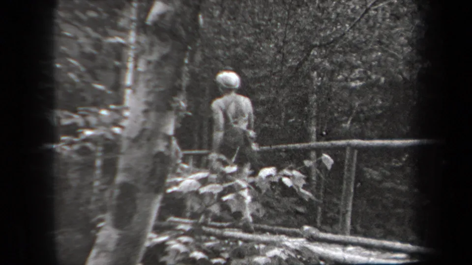 1938 New York home-movie frame with a woman in a hat near a bridge rail in a shaded forest setting.