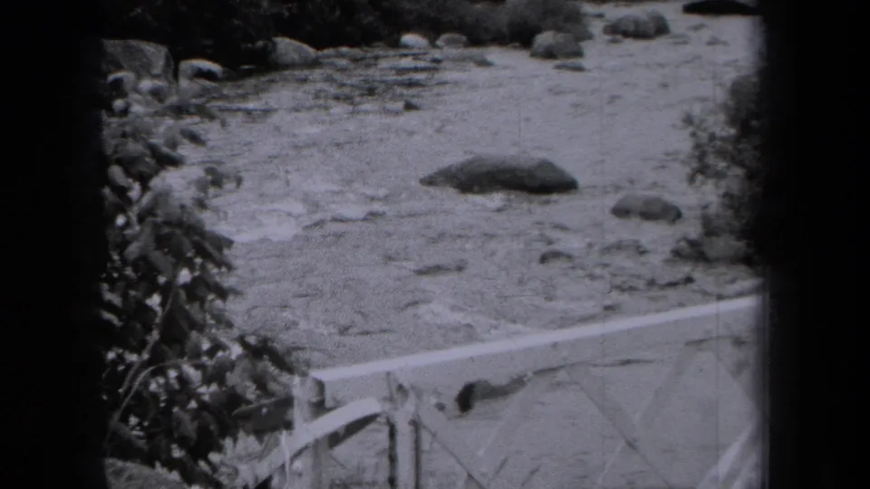 1938 New York 8mm frame showing shallow river water, scattered rocks, wooded edges, and part of a bridge railing.