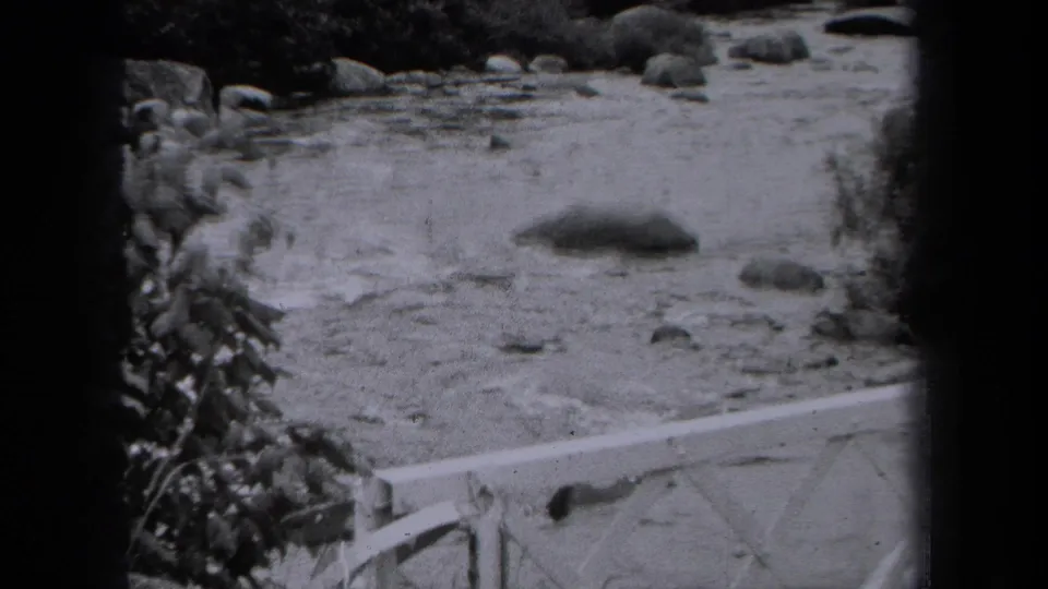 1938 New York 8mm frame showing shallow stream water, rounded rocks, foliage, and a pale railing or bridge edge.