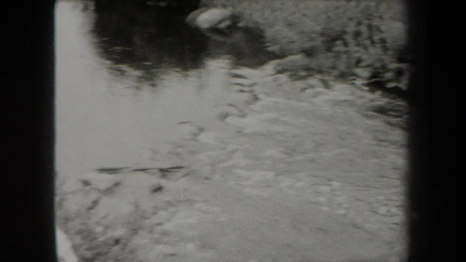 1938 New York 8mm frame showing a gentle stream with soft ripples, small rocks, and blurred vegetation along the water's edge.