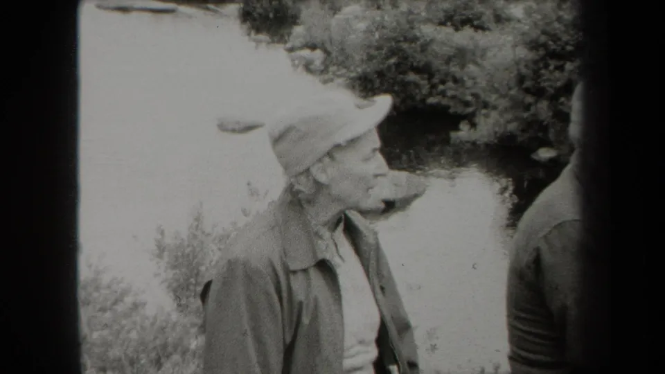 Black-and-white 1938 New York home-movie frame of a woman in a cap and jacket standing by a wooded creek with another adult partly visible.