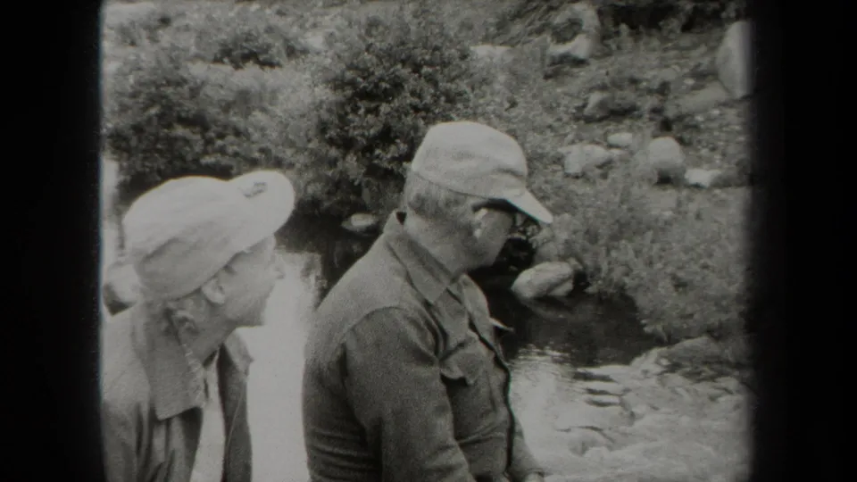 Black-and-white 1938 New York home-movie frame of a woman and man in caps standing near a rocky creek and dense brush.