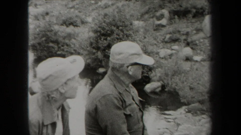 1938 New York 8mm frame of an older couple in hats standing outdoors near a small rocky creek.
