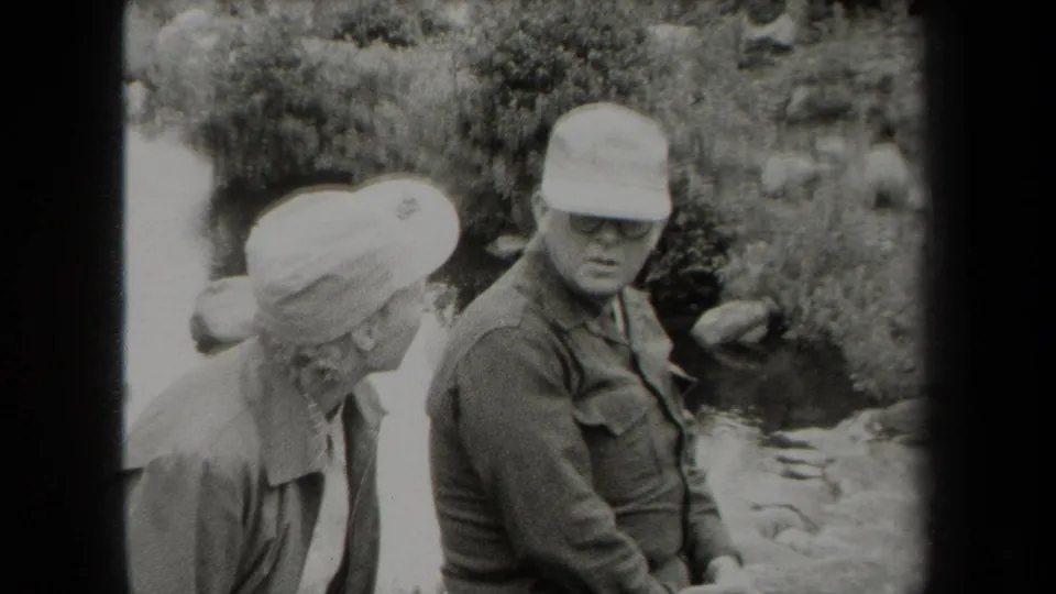 Older man in a cap and glasses turns toward an older woman in a light hat beside a New York creek in 1938 home-movie footage.