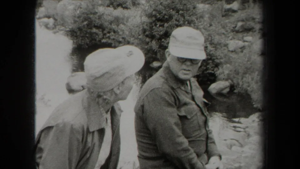 In a 1938 New York outdoor home movie, an older woman leans toward a capped man with glasses near a creek.
