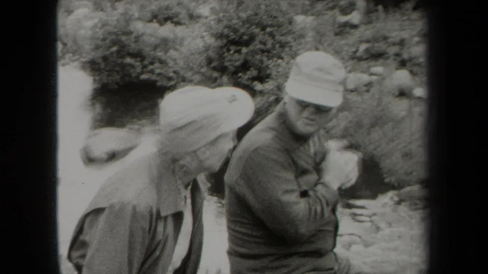 Older man in cap and glasses raises a hand as he talks beside an older woman near a rocky New York stream in 1938 footage.