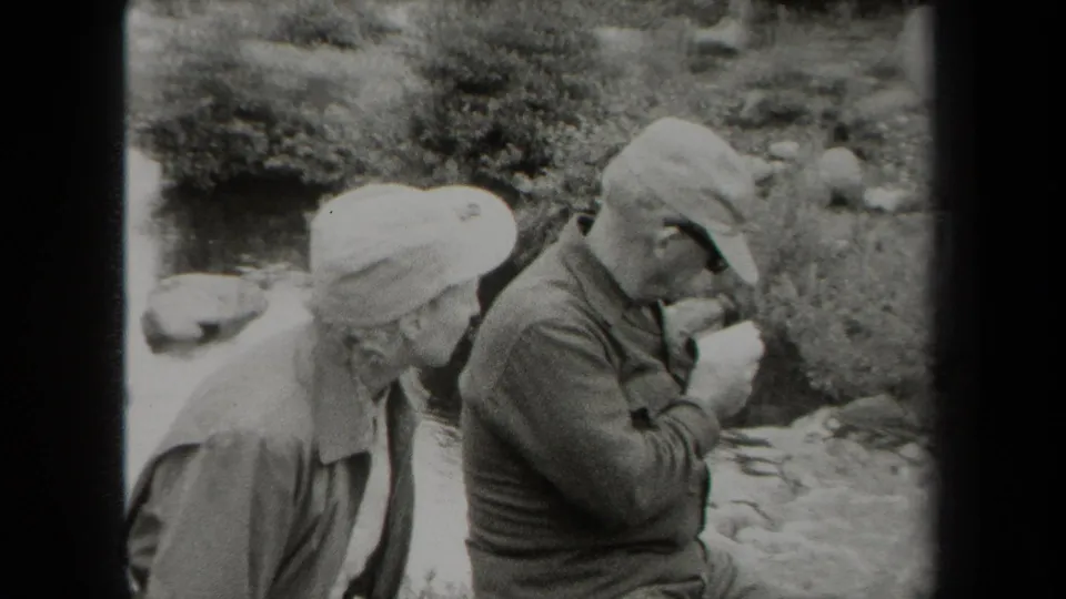 In metadata-supported 1938 New York, two uniformed men sit near a creek as one studies a folded paper or map.