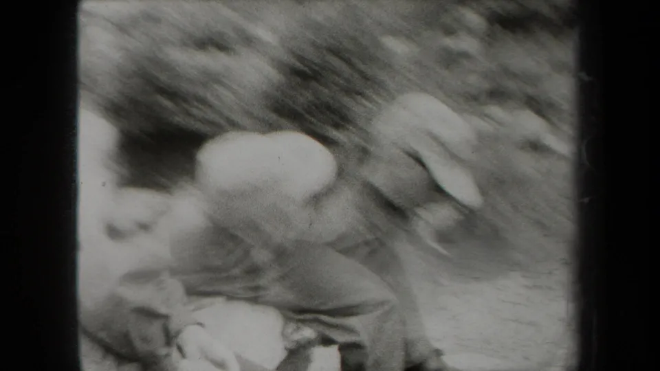 A blurred handheld frame shows two uniformed men beside a rocky creek in the supported New York home-movie setting.