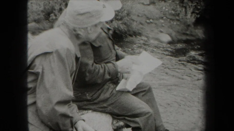 Two uniformed men sit at the creek edge in the 1938 New York clip, leaning toward a folded paper or map.