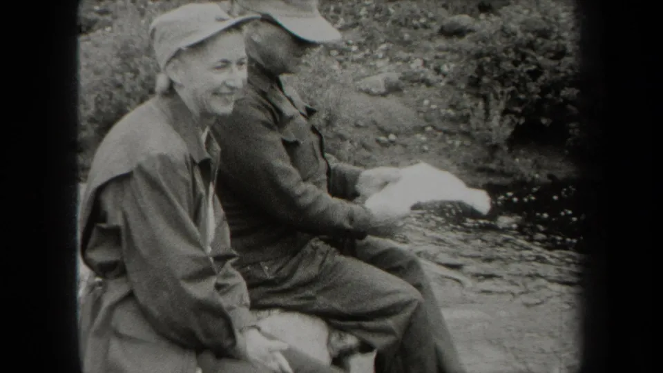 One uniformed man smiles toward the camera as his companion holds a paper near the creek in metadata-supported 1938 New York.