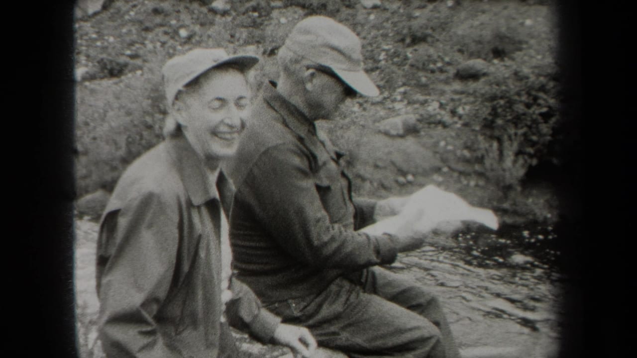 In metadata-supported 1938 New York footage, two men in caps sit by a rocky riverbank as one smiles toward the camera.