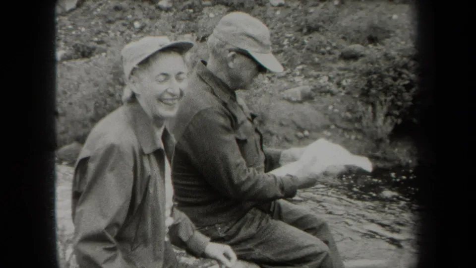 In metadata-supported 1938 New York footage, two men in caps sit by a rocky riverbank as one smiles toward the camera.