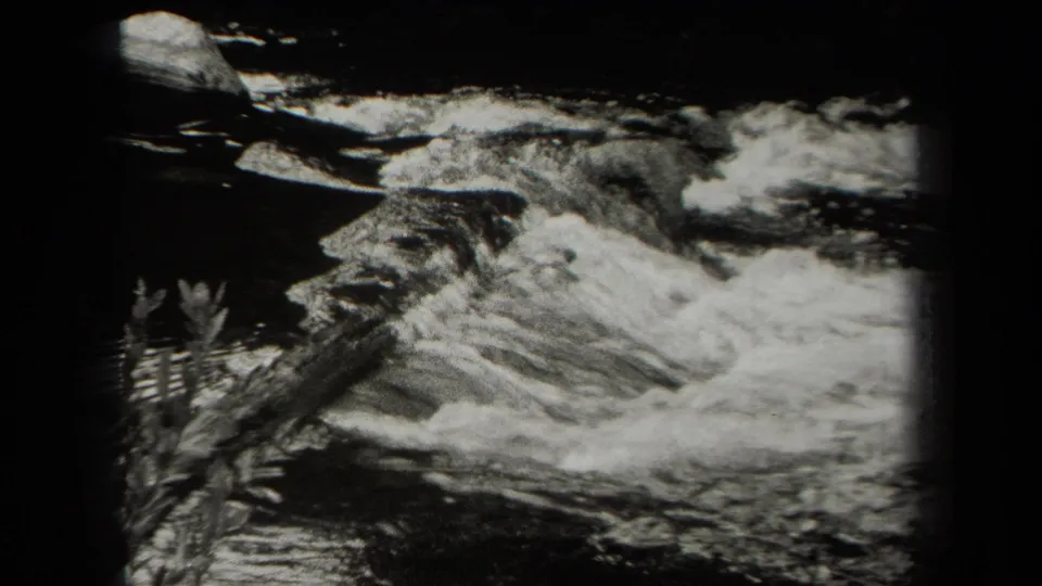 1938 New York 8mm frame showing rushing river water, dark rocks, and nearby leaves in soft black-and-white focus.