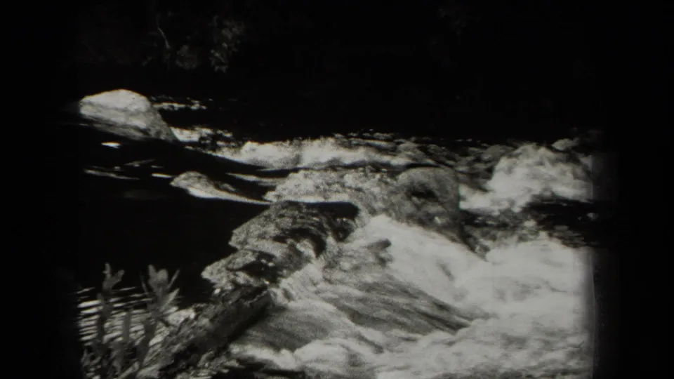 Black-and-white 8mm view of a New York river current with white water, rocks, and plants along the edge.