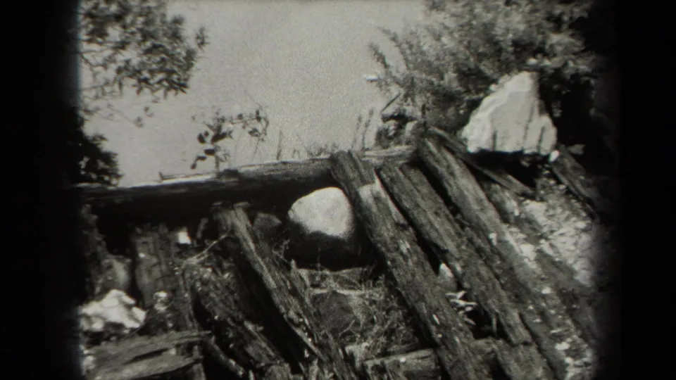 Black-and-white New York home-movie frame showing weathered planks, stones, brush, and bright sky around a damaged outdoor structure.