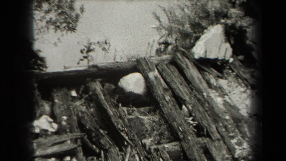 1938 black-and-white home-movie frame of a collapsed or damaged wooden structure with stones, debris, brush, and dark frame borders.