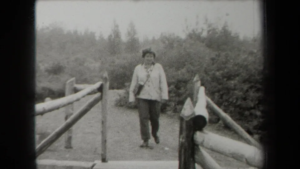 In 1938 New York home-movie footage, an adult woman in a light jacket and dark pants approaches on a wooden bridge in a wooded area.