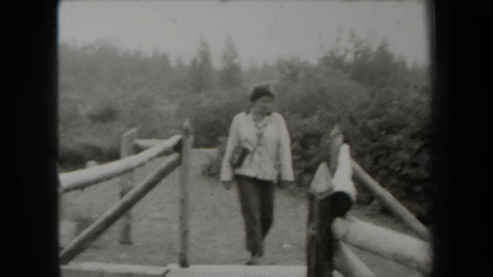 The woman moves closer to the camera on the rustic bridge, with log railings and dense foliage visible in the 1938 New York footage.