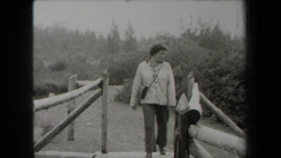 Closer in frame, the adult woman appears to glance sideways while walking on the wooden bridge in a wooded New York setting.