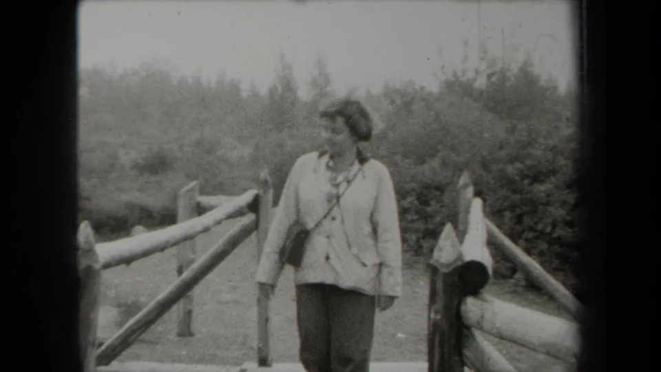 In metadata-supported 1938 New York footage, a woman stands near a rustic wooden fence in a wooded or landscaped area.
