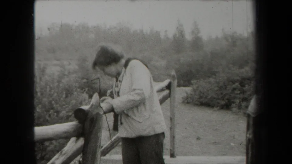 A woman in a light jacket leans toward the wooden fence, with dense vegetation and an open path visible behind her.