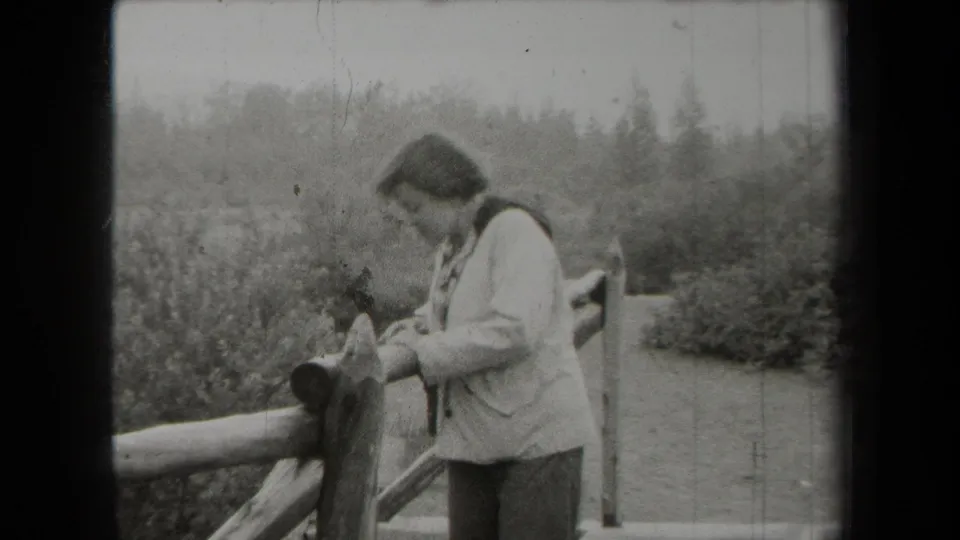 The final frame shows the woman beside the fence in the 1938 home-movie clip, surrounded by shrubs, trees, and a narrow path.