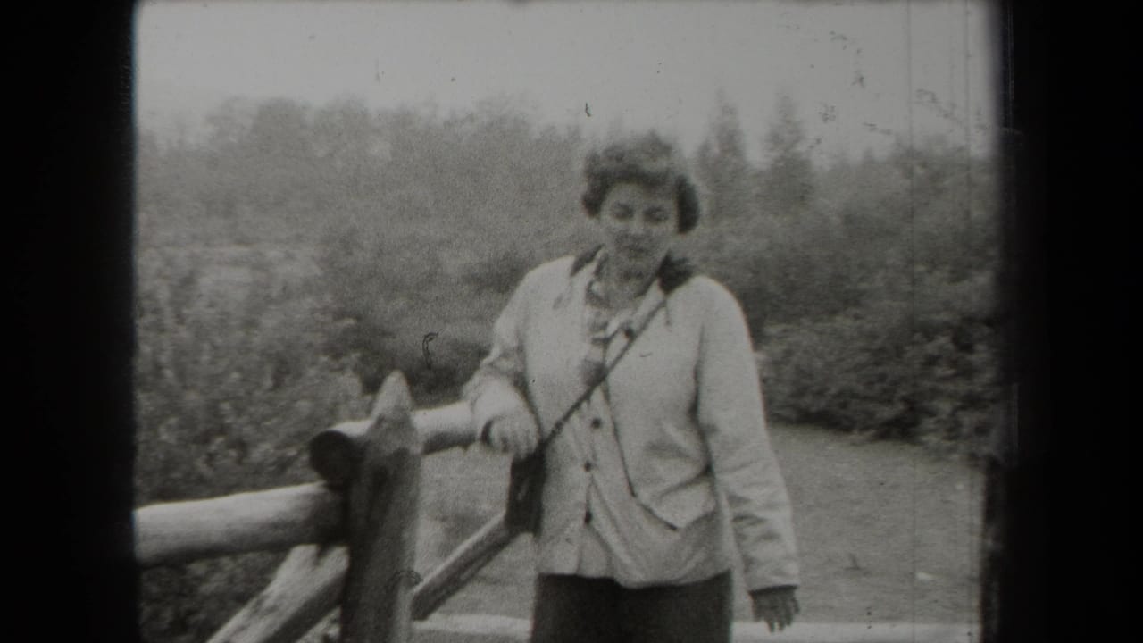 Adult woman in a light jacket stands beside a rustic wooden fence in a rural setting.