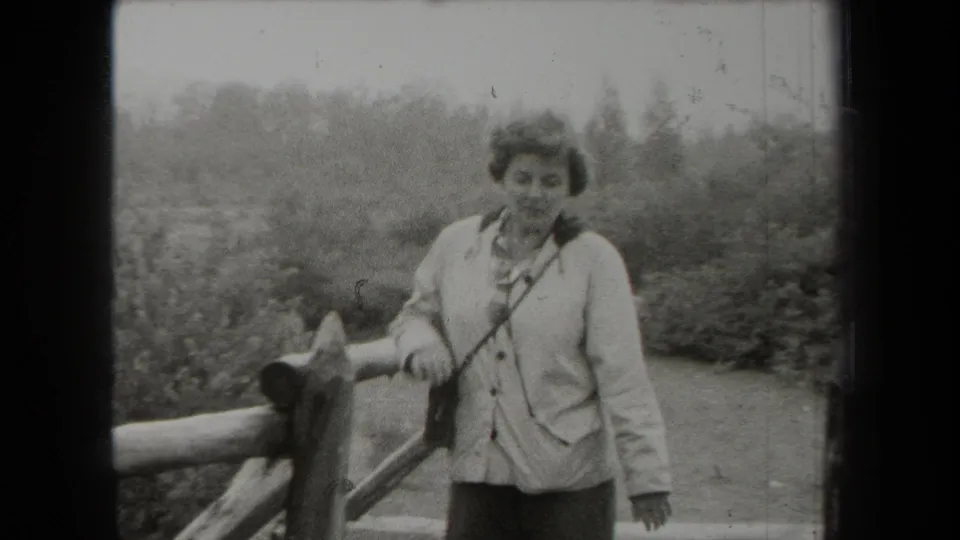 In 1938 New York home-movie footage, an adult woman stands near a wooden fence with trees and brush behind her.