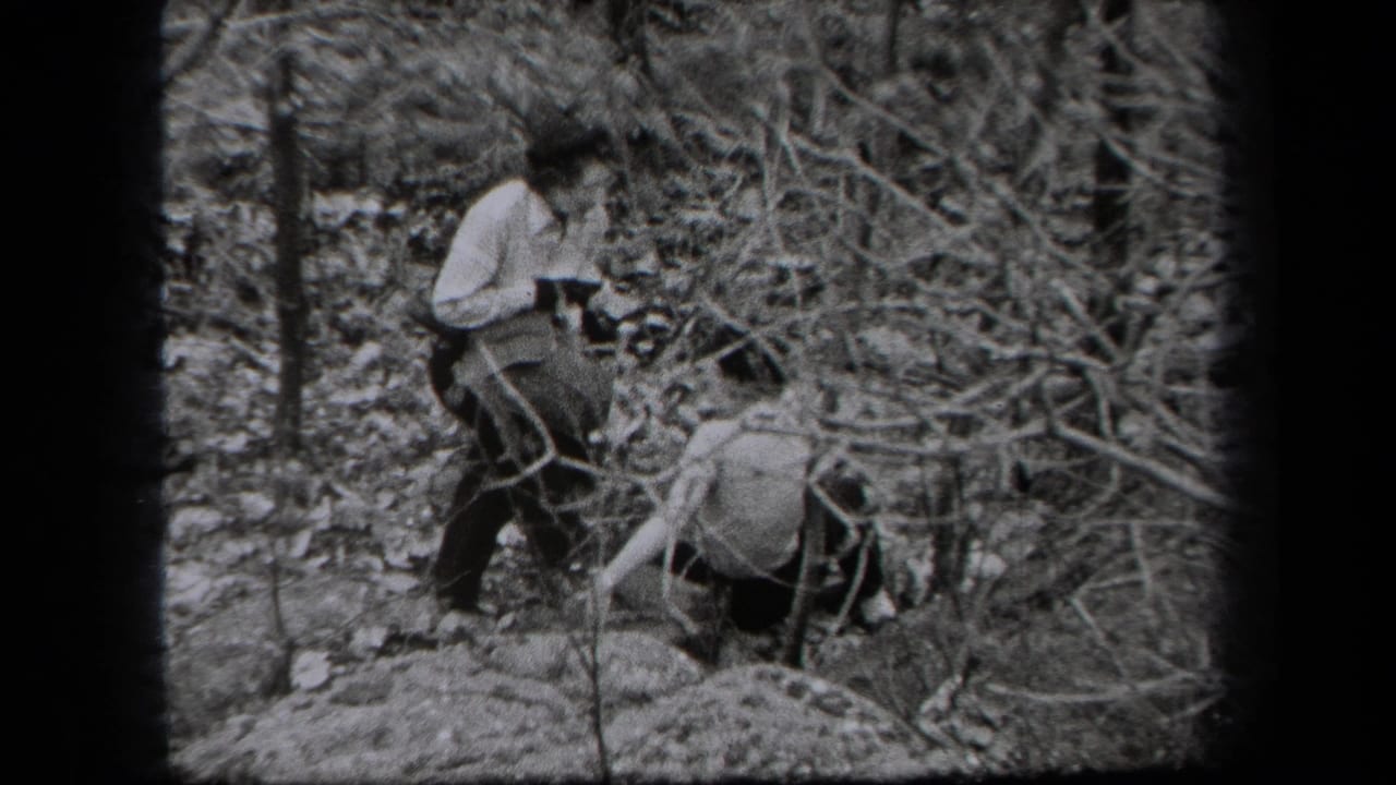 1938 New York home-movie frame showing two adults navigating a wooded area with leaf litter and bare branches.