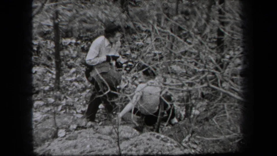 1938 New York home-movie frame showing two adults navigating a wooded area with leaf litter and bare branches.