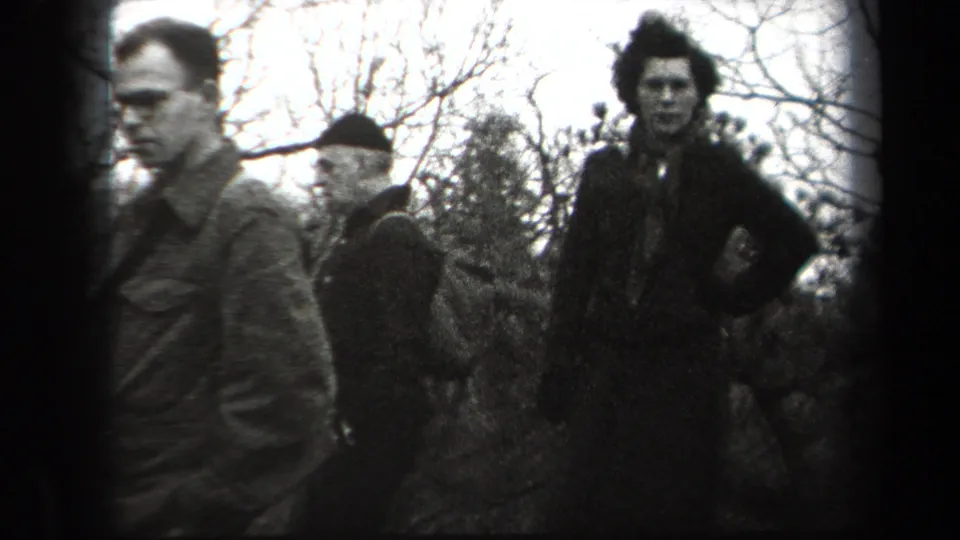 1938 New York home-movie frame showing three adults in period outerwear near bare trees, including one person in a dark coat and hat.