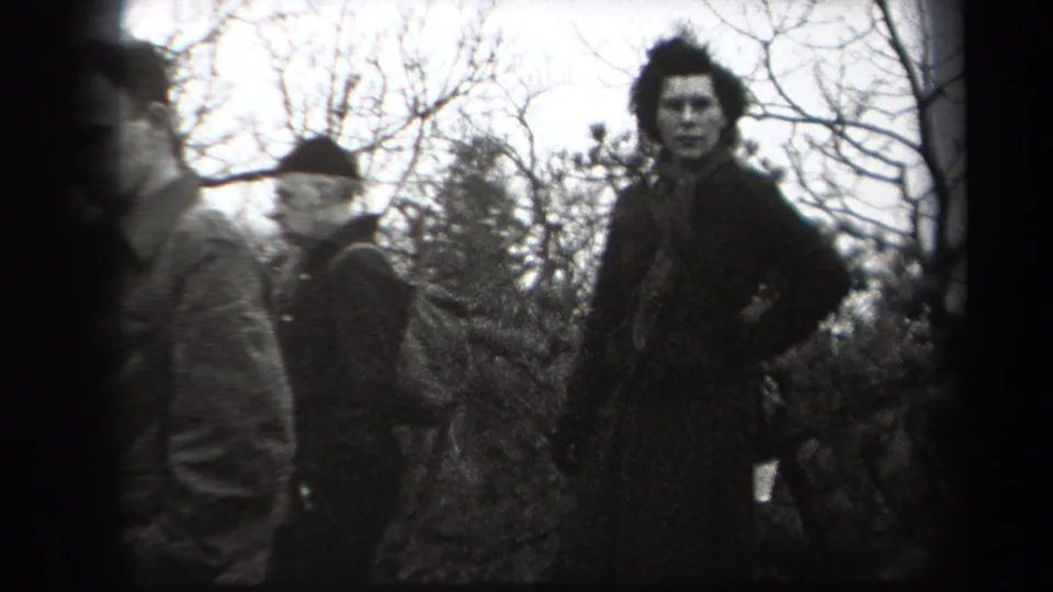 In 1938 New York home-movie footage, adult men in coats and hats gather in a wooded outdoor area while one man looks toward the camera.