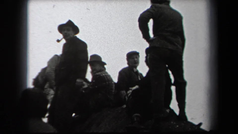 Adult men in period coats and hats sit and stand on a rocky outcrop, with a pipe-smoking figure visible in the foreground.