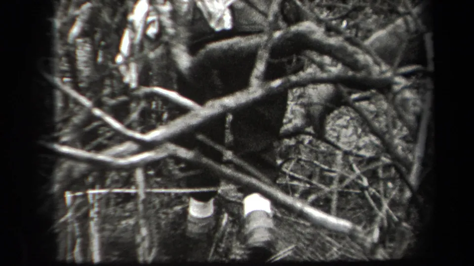Adult man viewed from behind in a wooded area, framed by branches and wearing dark outdoor clothing with light socks or gaiters.