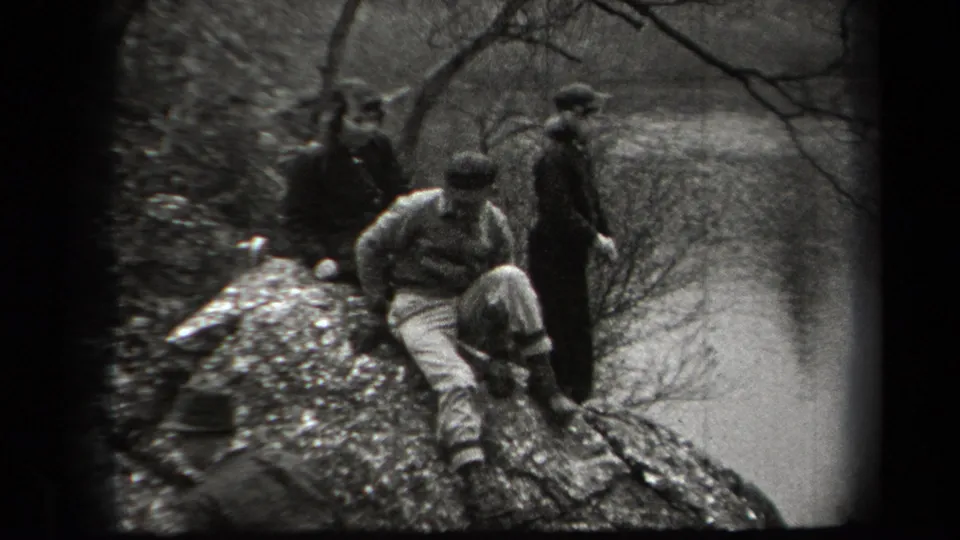 Young men in caps and casual clothing sit and stand on a rocky outcrop in a wooded New York setting, filmed in black-and-white 8mm.