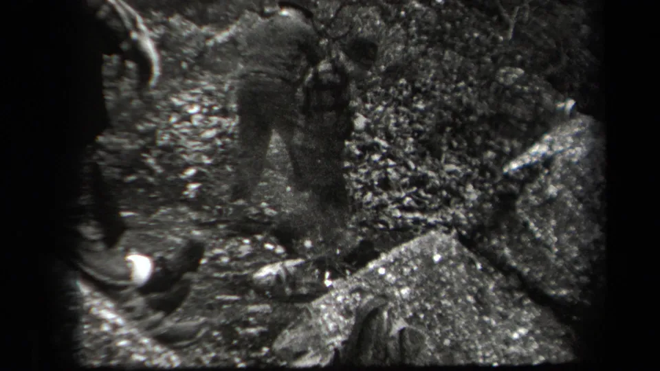 Men in 1930s casual clothing stand close together among rocks and leaf litter in a wooded New York outdoor scene.