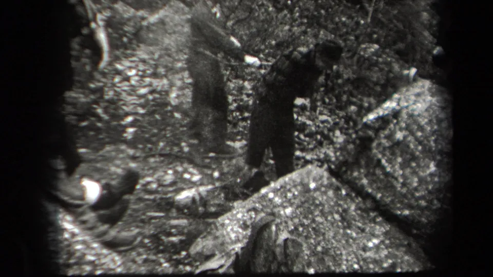 Two young men stand beside rocks and leaves in the wooded 1938 New York home-movie scene, with another figure partly cropped at left.