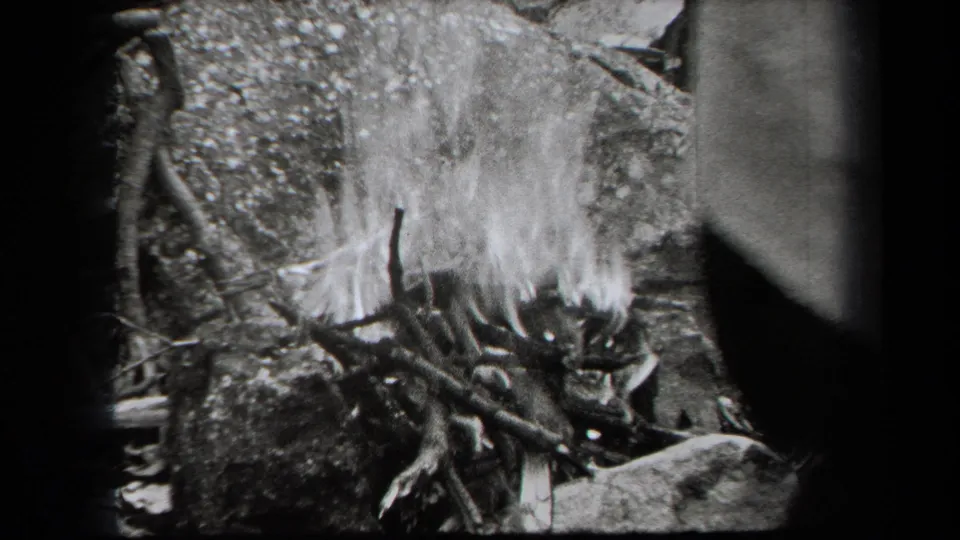 1938 New York 8mm frame showing a small campfire with sticks, flame, smoke, rocks, and dark woodland edges.