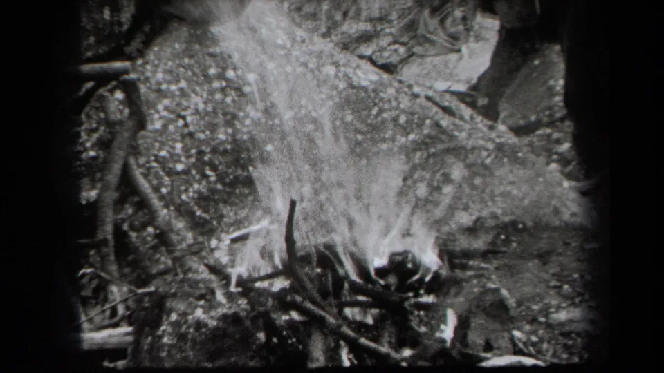 1938 New York 8mm view of flames rising from a rough stack of sticks against rocks and forest debris.