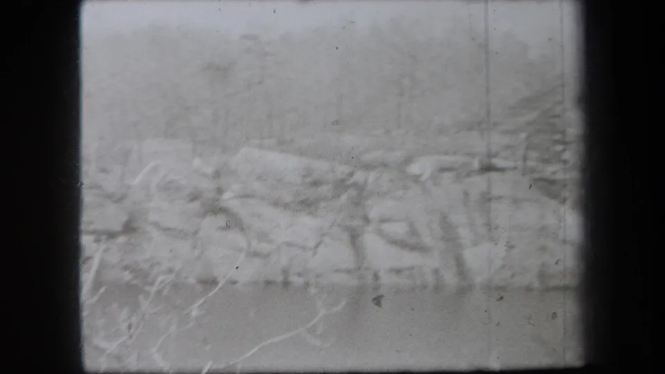 1938 New York home-movie frame with a pale snowy bank, leafless brush, rocks, and water under soft winter haze.