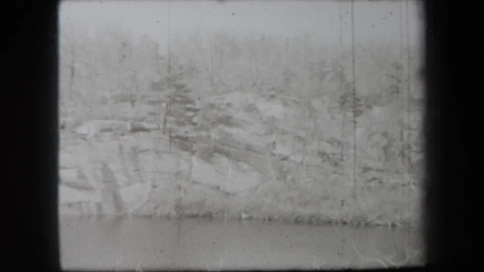 1938 New York 8mm frame showing a soft-focus winter shoreline, scattered rocks, bare trees, and a narrow strip of water.