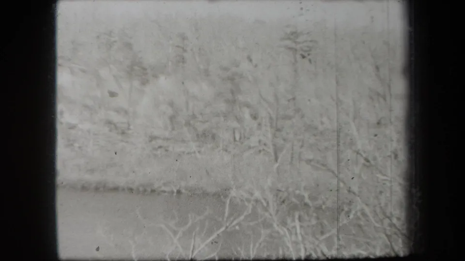 1938 New York winter frame with leafless foreground branches, a snow-covered bank, wooded terrain, and calm water.