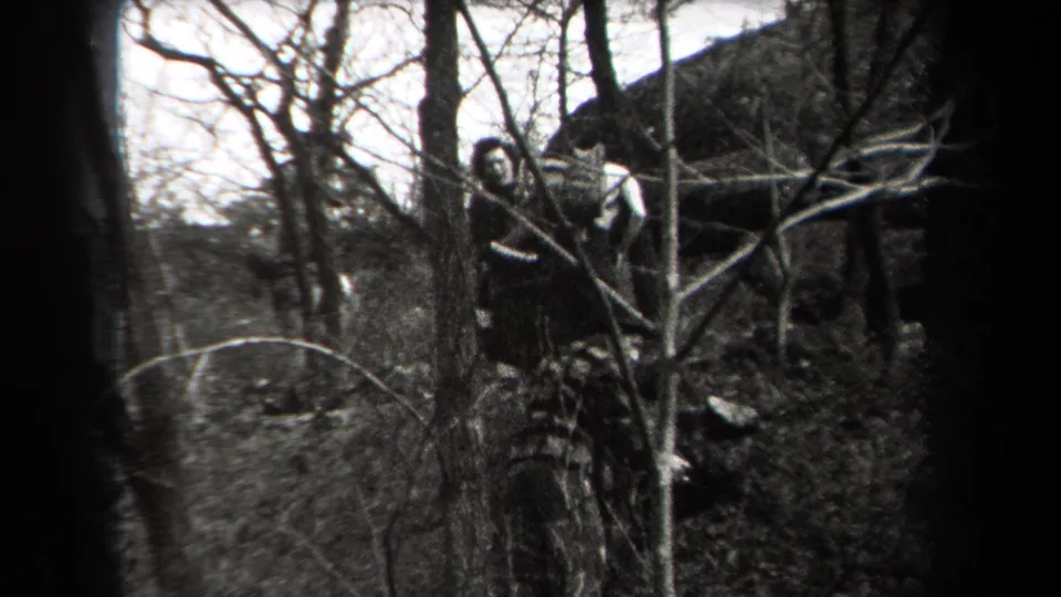 A man stands partly obscured by tree trunks and bare branches on rocky forest ground in the 1938 New York home-movie scene.