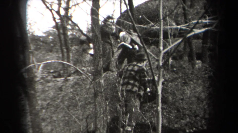 A hiker in period outdoor clothing moves through brush and bare branches in the 1938 New York wooded scene.