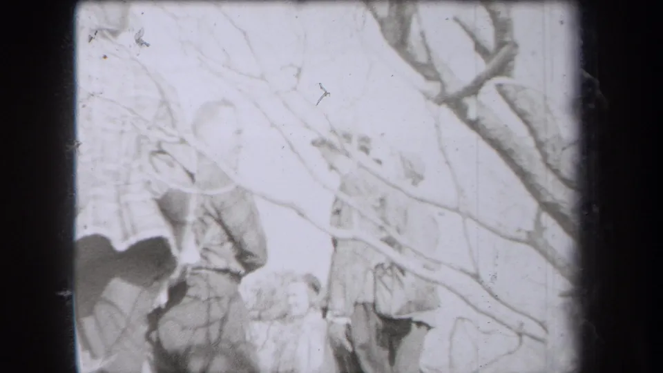 Overexposed 8mm footage shows people in hats and coats partly obscured by foreground branches in a New York woods outing.