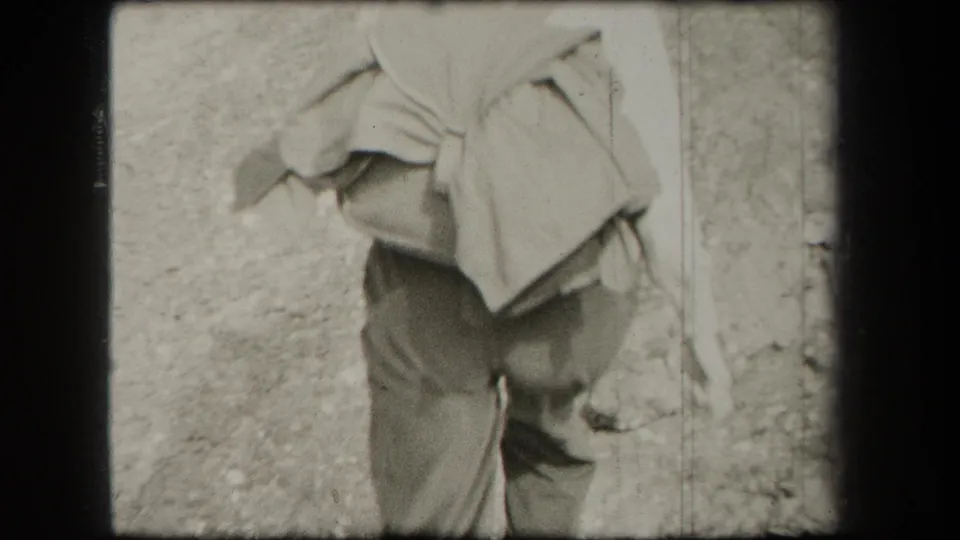 A woman in period outdoor clothing carries a large pack or blanket roll while walking through wooded terrain in New York, 1938.